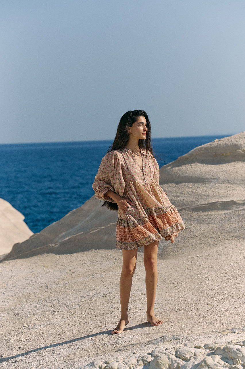 Full-length view of woman on white coastal rock by deep blue sea wearing the Daughters of India Bhoomi Mini Dress in Sunset, showing the complete boho silhouette