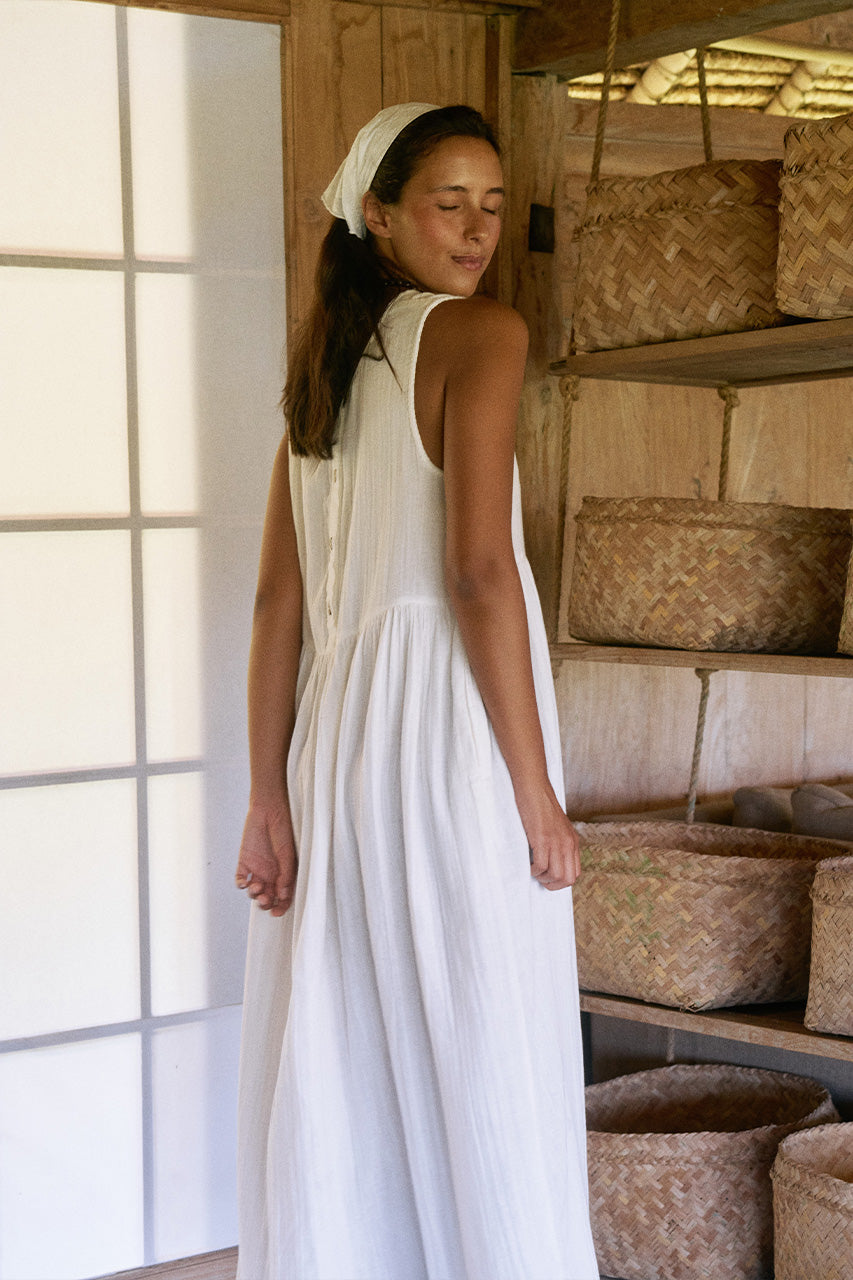 Nadi Dress White — back view showing scooped back neckline, white double gauze cotton midi dress with flowing gathered skirt, rustic bamboo interior with woven baskets | Daughters of India