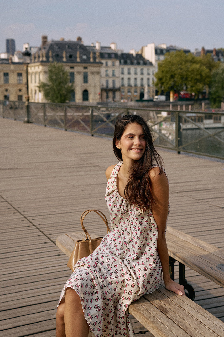 Smiling woman seated on a wooden bench by the River Seine in Paris wearing the Daughters of India Sundress Midi in Peony, with the Parisian cityscape in the background
